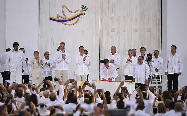 Gente vestida de blanco en la audiencia y el escenario incluyendo la Jefa de Estado durante la ceremonia de la firma de paz entre el gobierno de Colombia y las FARC. Al fondo hay una imagen de una paloma con un laurel.